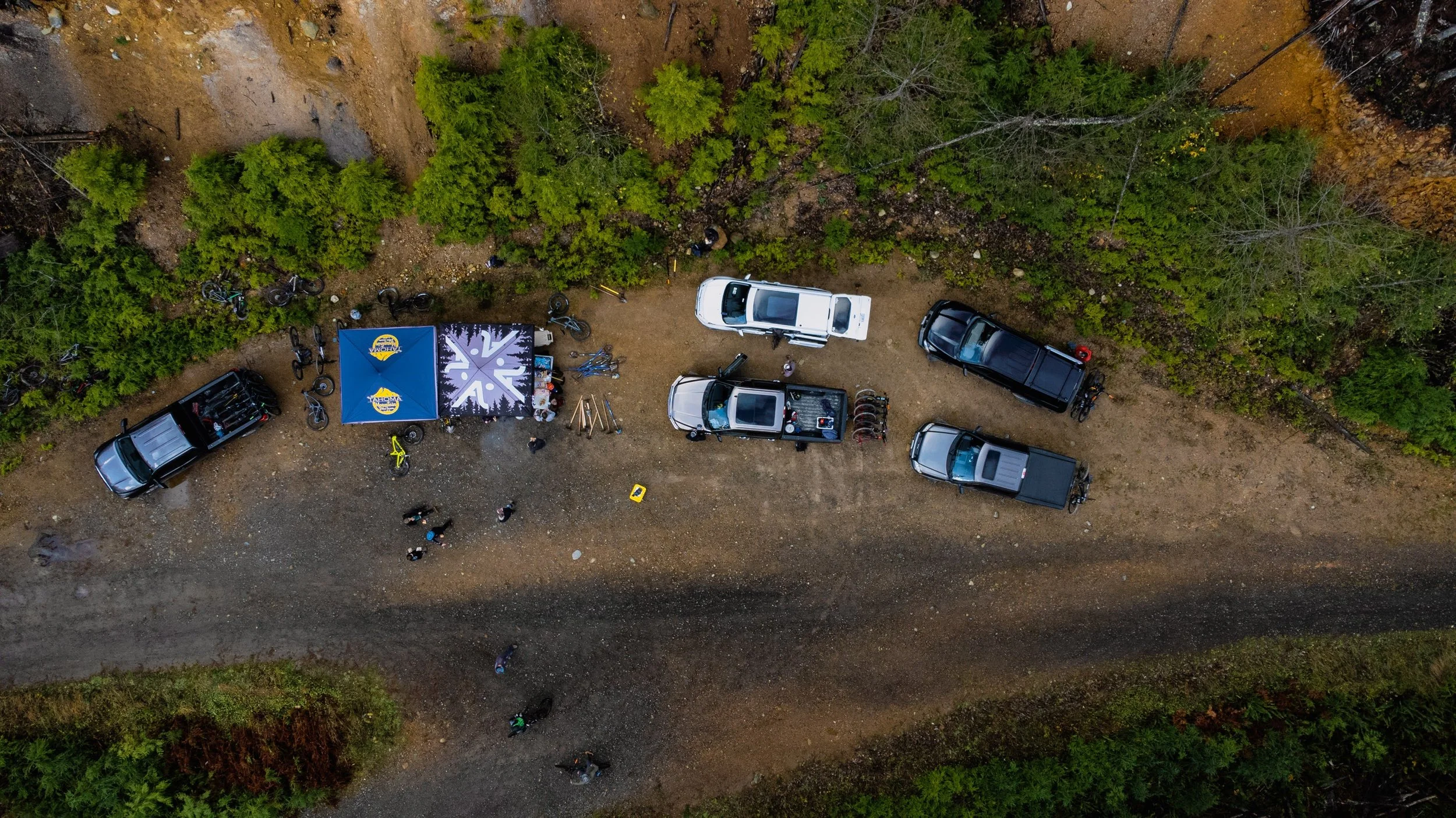 An aerial view of a dirt area with six parked vehicles, a tent, and several people gathered around