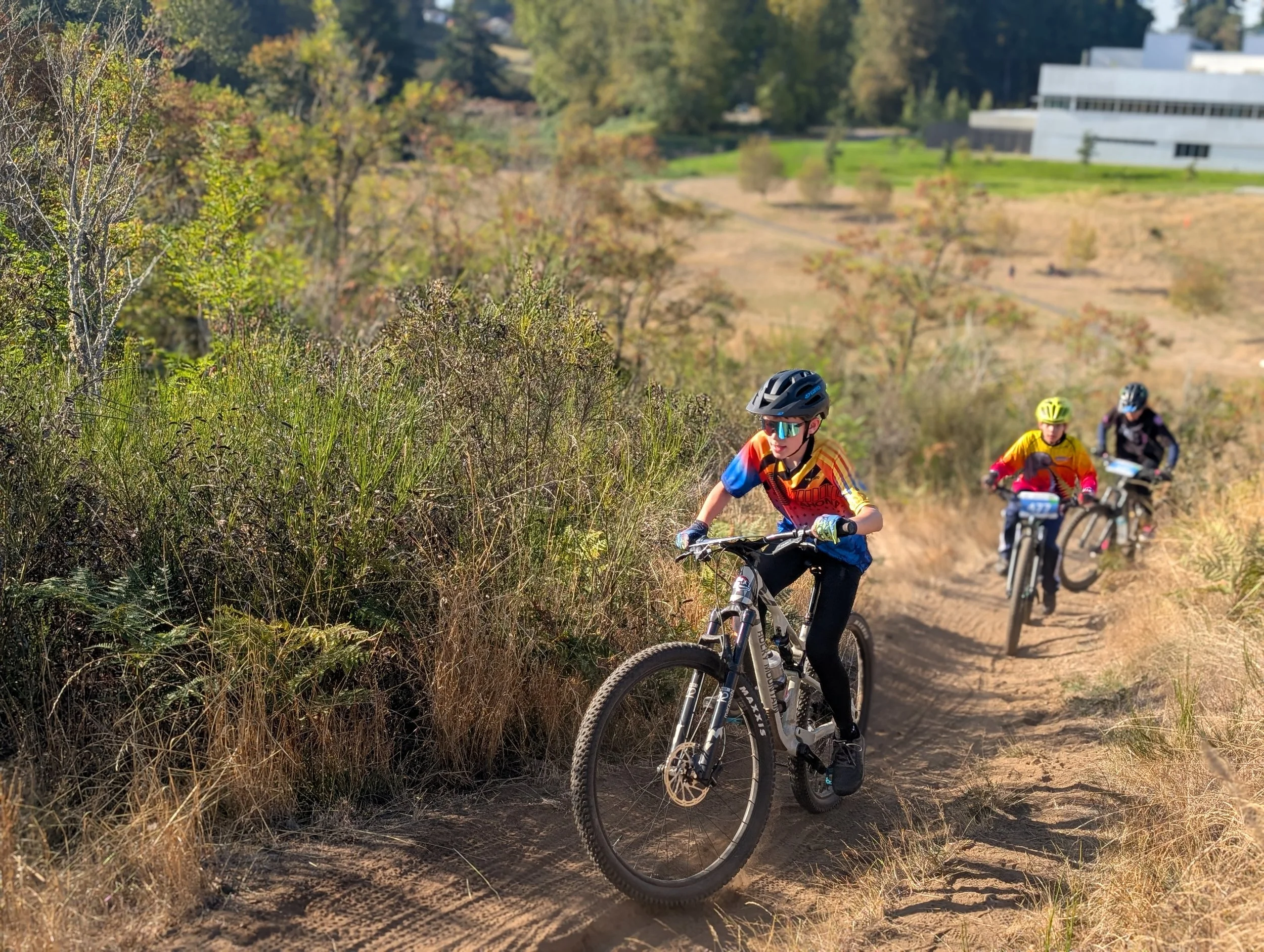 Three children mountain biking on a dirt trail surrounded by dry grass and bushes, with trees and buildings in the background.