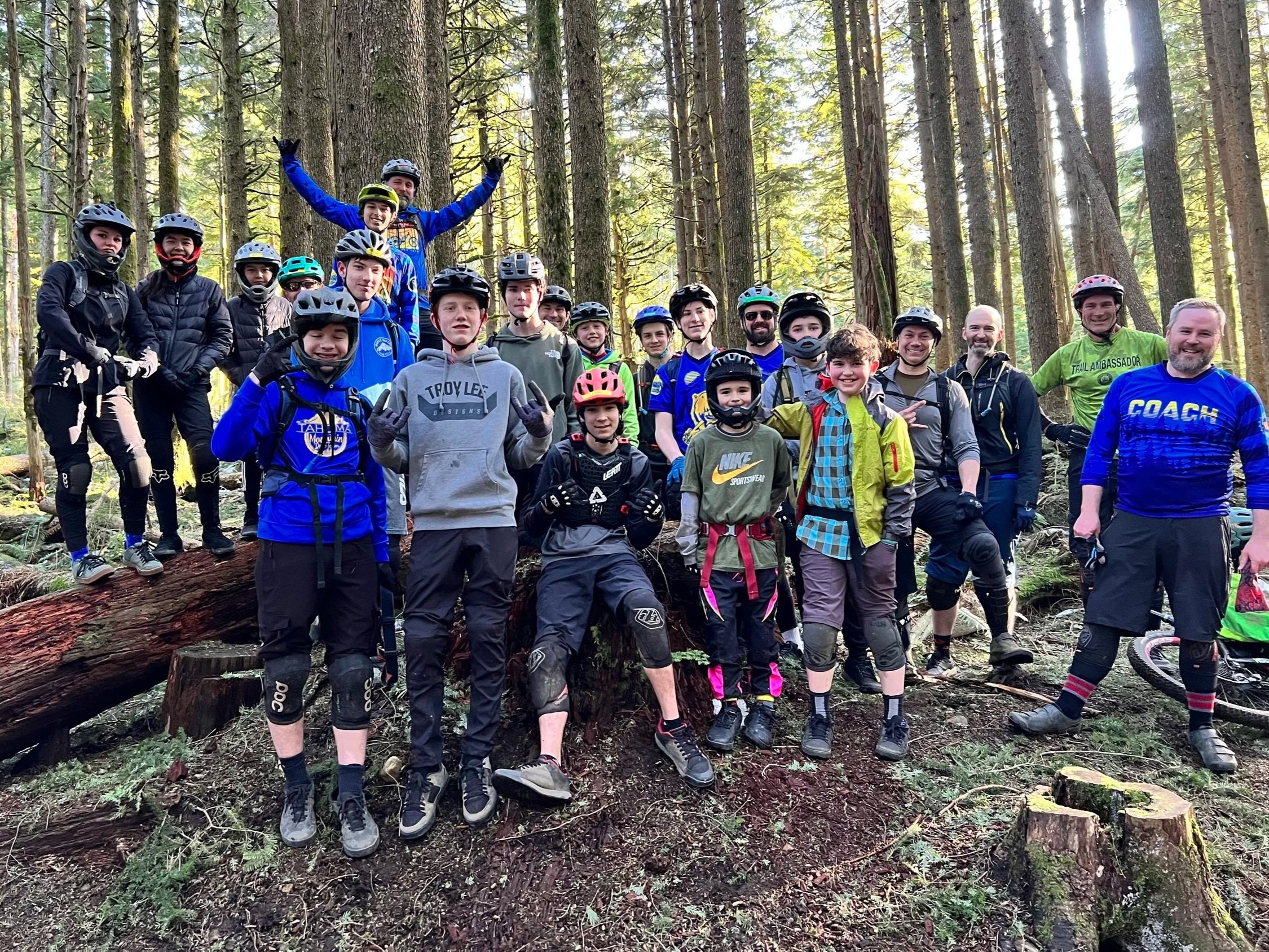 A group of young mountain bikers and their coaches posing together in a wooded forest, all wearing helmets and biking gear.
