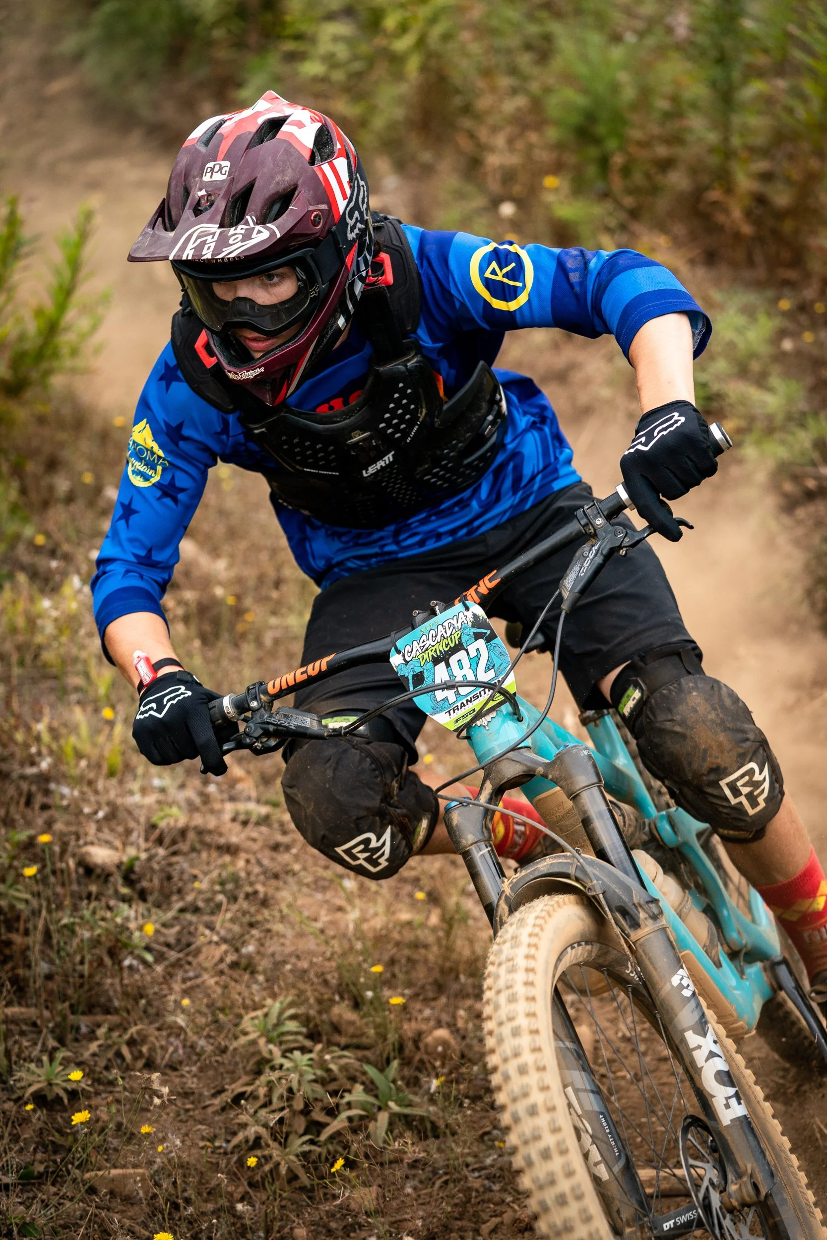 A mountain biker wearing a full face helmet, blue jersey, and gloves riding downhill on a dirt trail.