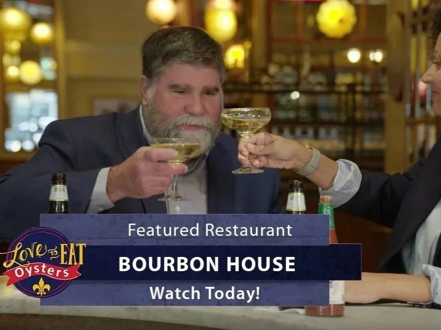 Two people with gray hair and beards at a restaurant bar, clinking champagne glasses, with bottles on the bar, in a warmly lit setting. A promotional overlay for Bourbon House restaurant, featuring the Love to Eat Oysters logo, with text endorsing it as a featured restaurant.