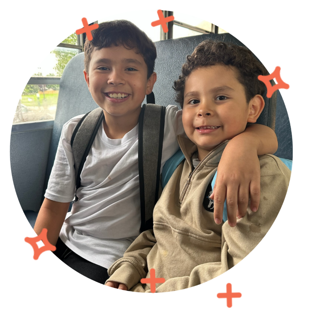 Two young boys sitting next to each other on a school bus, smiling. The older boy has his arm around the younger boy, who has curly hair and is wearing a beige hoodie.