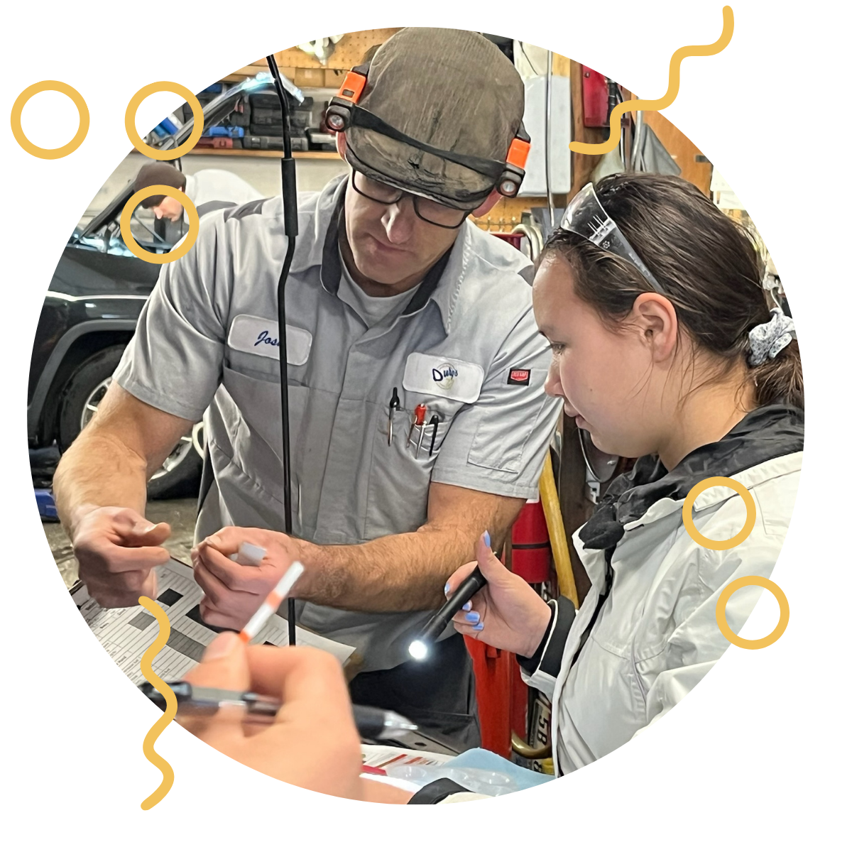 Two people with safety glasses and work uniforms engaged in a task in a workshop, with tools and equipment in the background.