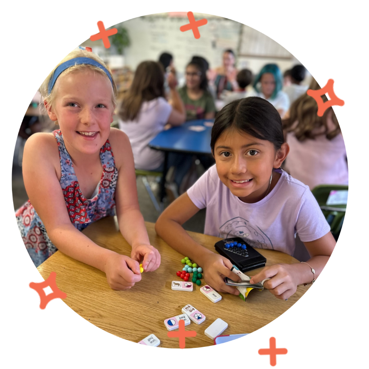 Two young girls playing a game with tiles and colorful game pieces at a table in a classroom setting, with other children sitting at tables in the background.