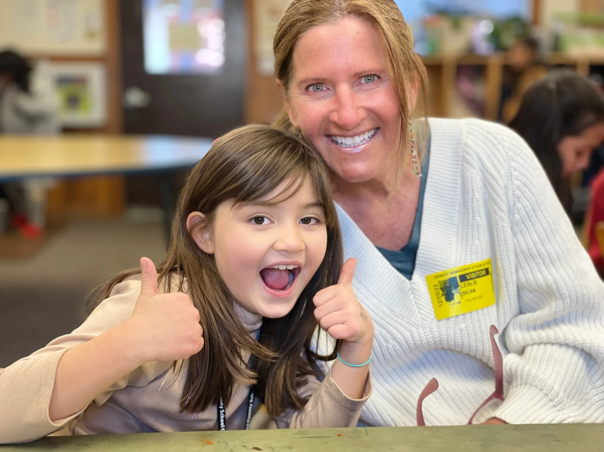 A smiling woman and girl posing together, giving thumbs up in a classroom.
