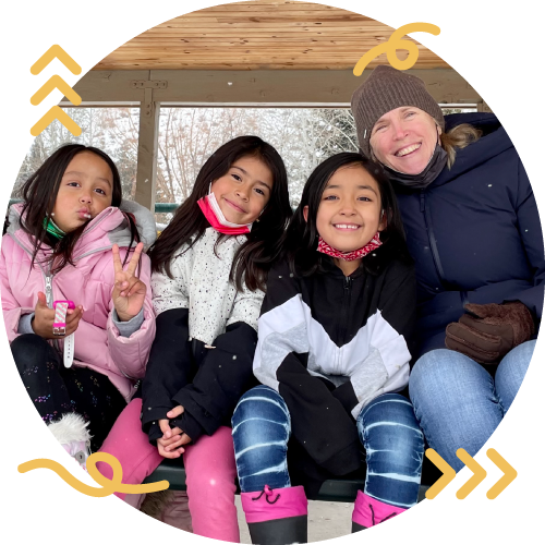 Four females sitting together outdoors on a wooden structure, smiling and making peace signs. One is wearing a pink jacket, one in a white and black jacket, and another in a black and white jacket. The adult woman is wearing a brown beanie and blue jacket.