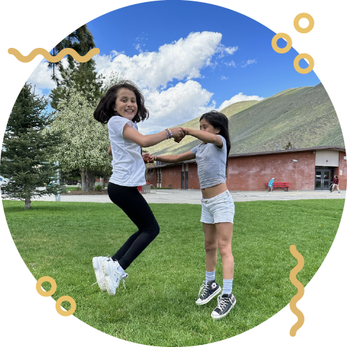 Two young girls playing and holding hands in a grassy park with a building, trees, and a mountain in the background on a sunny day.