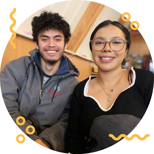 A smiling young man with curly hair and a young woman wearing glasses, sitting indoors with warm lighting and wooden decor in the background.