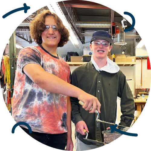 Two young men smiling and standing in a workshop. One is wearing a colorful tie-dye t-shirt and sunglasses, and the other is wearing safety glasses, a dark cap, and a dark green jacket. They are holding a metal object, possibly a tool or a piece of equipment.