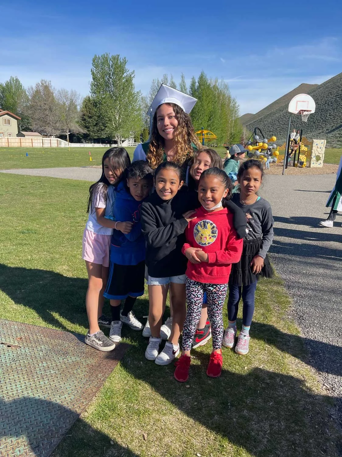 A woman in a graduation cap and gown with six children in a park on a sunny day.
