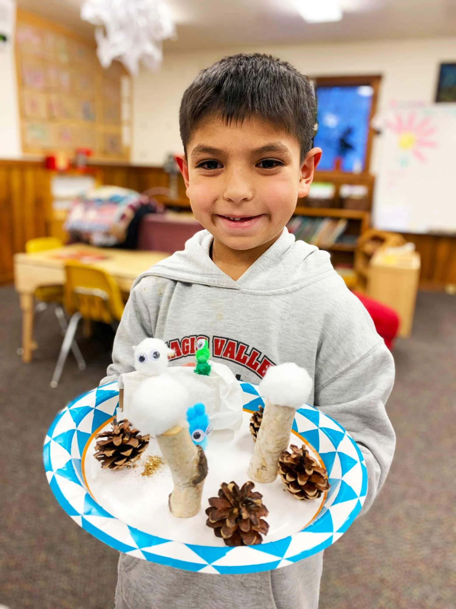 A young boy smiling and holding a tray with a craft project resembling a snowman, made with pinecones, cotton balls, pipe cleaners, and googly eyes, in a classroom setting.