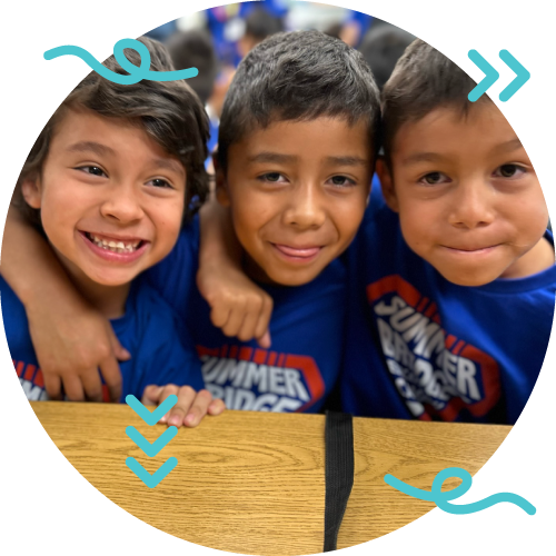 Three young boys wearing blue shirts with 'Summer Ridge' logo, smiling, with their arms around each other, standing behind a wooden table in a crowded indoor setting.