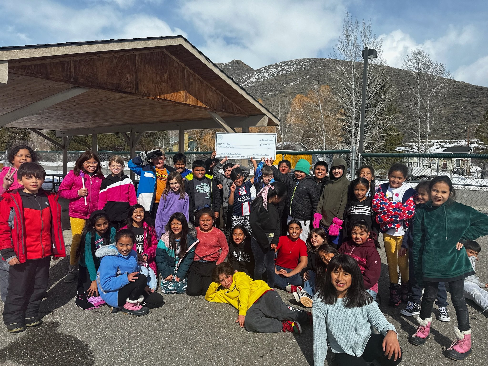 A group of children outdoors on a cloudy day, some holding a large check, standing under a wooden shelter near a fenced area with mountains in the background.