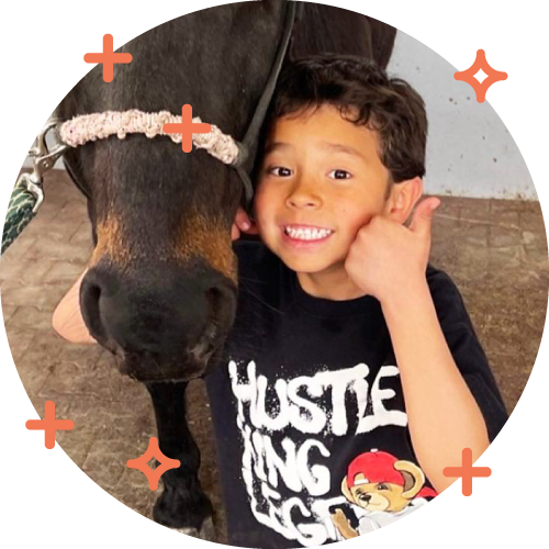 A young boy smiling and giving a thumbs-up next to a horse with a harness in a stable.