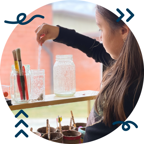 A young girl is adding salt to a glass jar with a spoon, on a window sill with paintbrushes in jars and planting pots nearby.