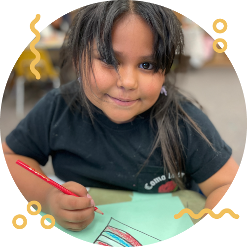 A young girl with long dark hair and a black shirt coloring at a table in a classroom.