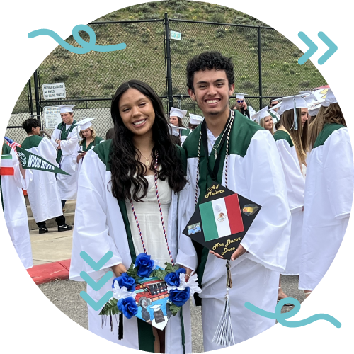 Two graduation students in caps and gowns smiling at the camera at an outdoor graduation ceremony.