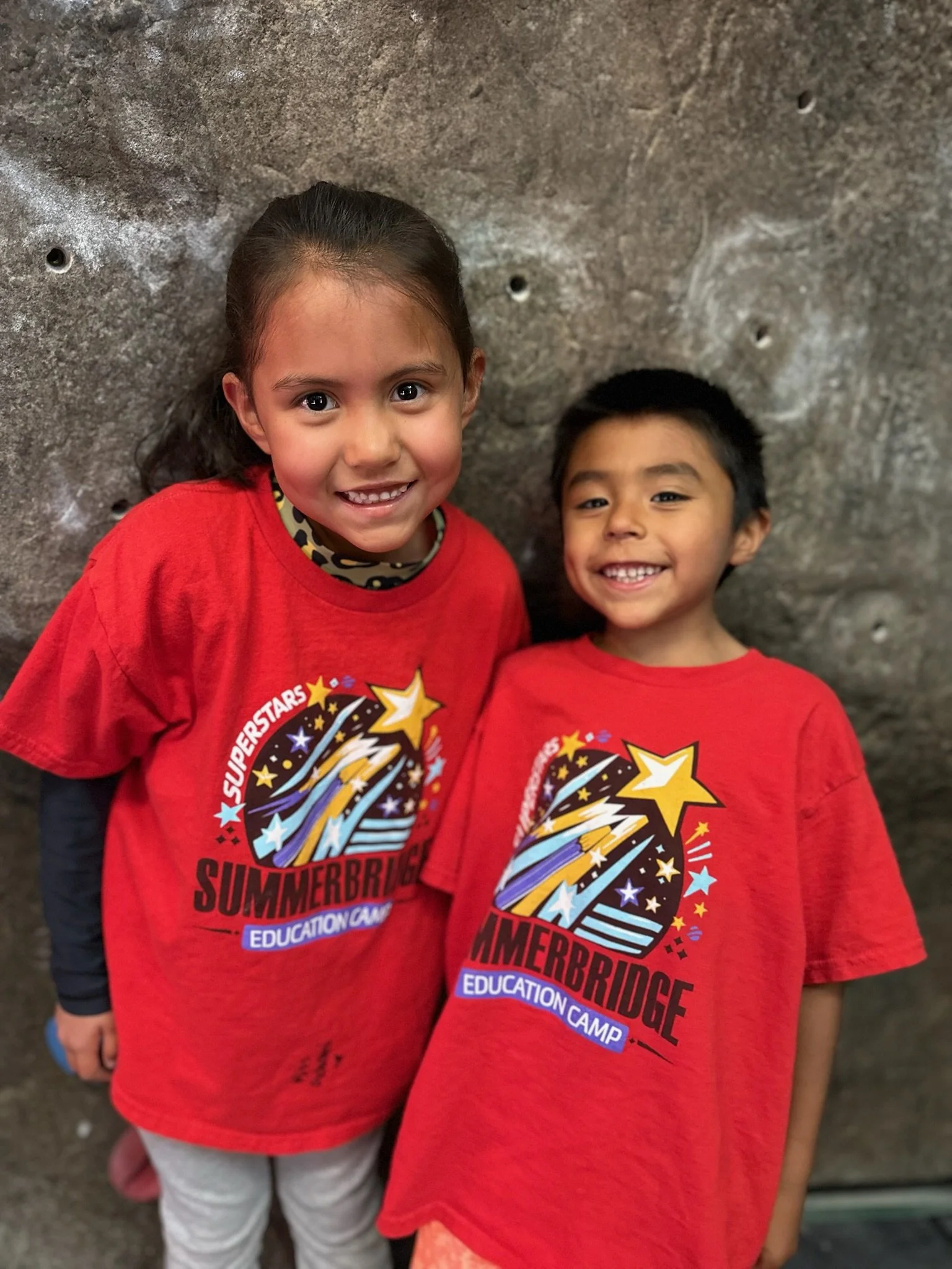 Two children wearing red 'Summerbridge Education Camp' T-shirts standing against a rock climbing wall, smiling at the camera.