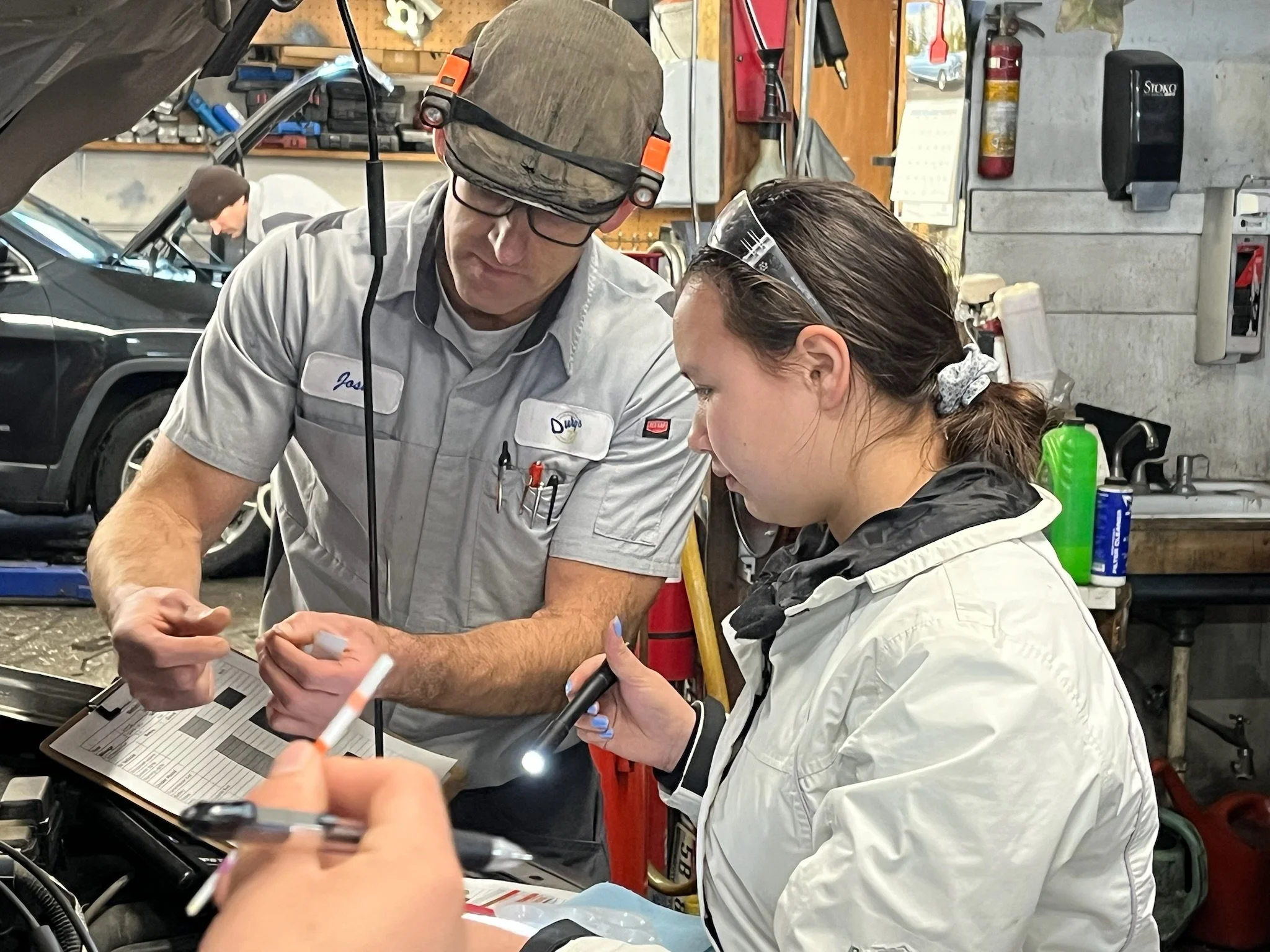 Two mechanics in a garage working together on a car, one holding a flashlight and the other reviewing a clipboard with tools and equipment in the background.