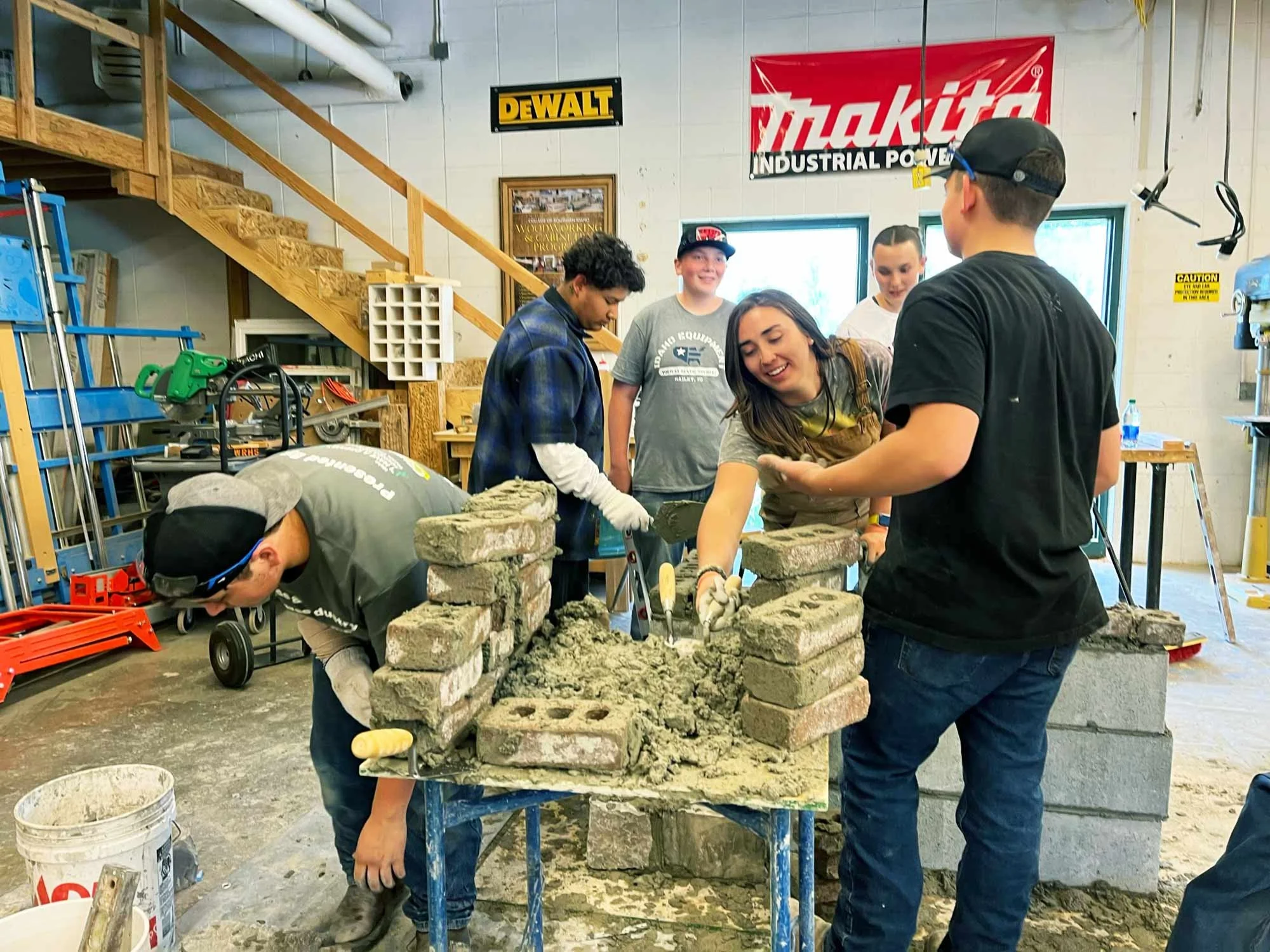 Group of young adults working together on a brick wall project inside a workshop. They are building with bricks and mortar, smiling and collaborating. There are tools and equipment around, with wood stairs and banners in the background.