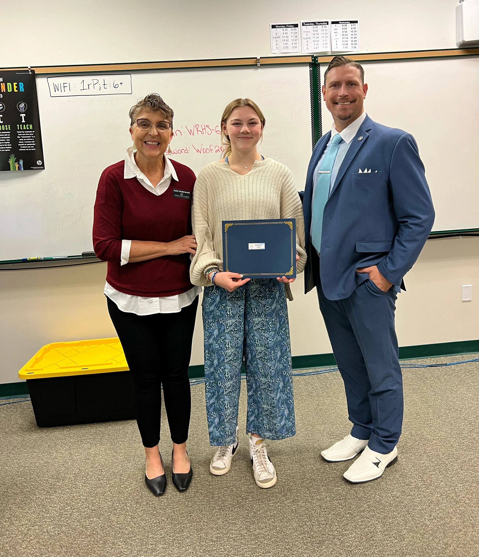 A young woman receiving an award or certificate from an older woman and a man in a blue suit in a classroom with a whiteboard.