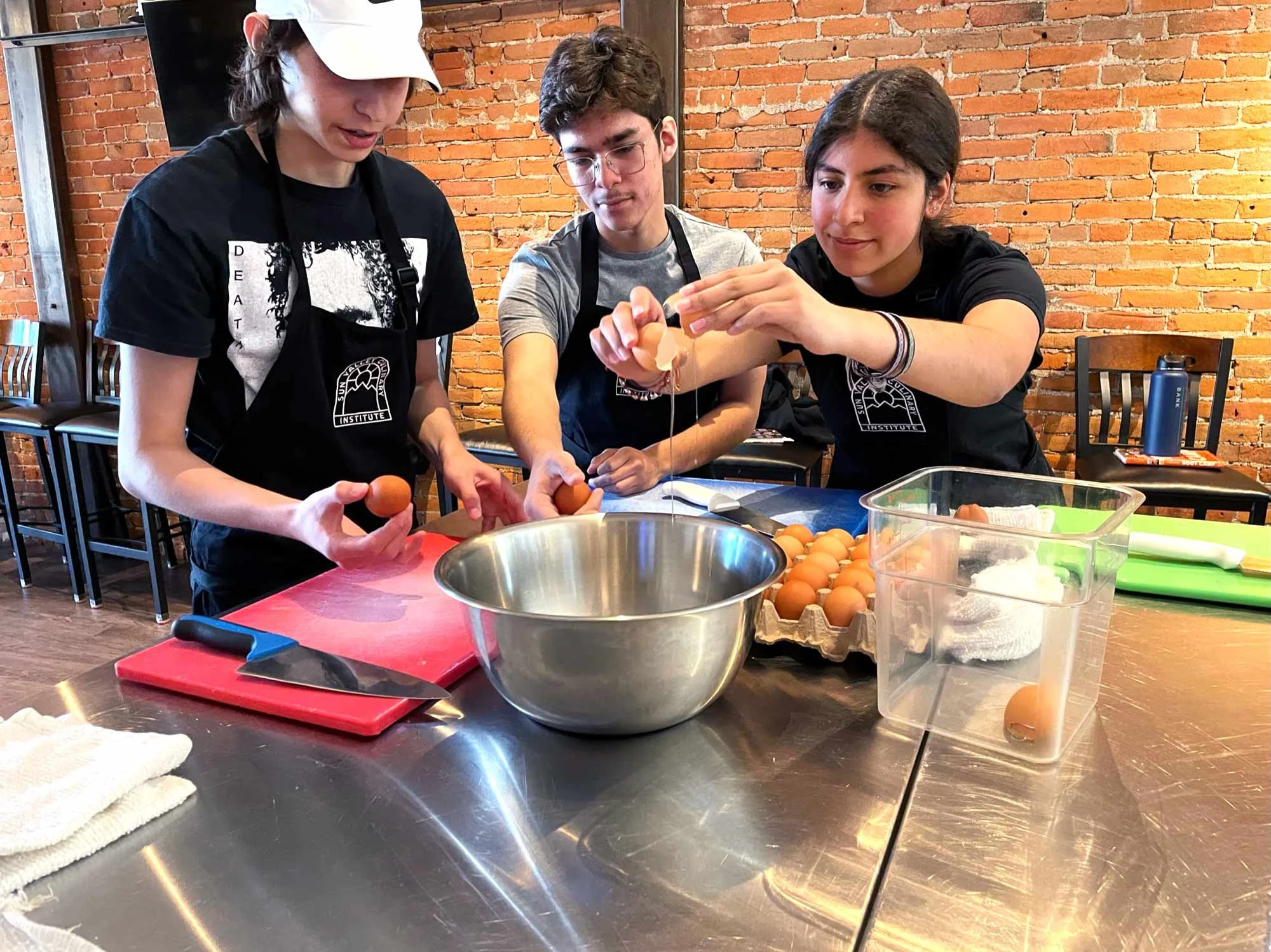 Three young people cracking eggs into a metal mixing bowl while standing around a kitchen counter with a box of eggs, a knife, and a container, in a room with exposed brick walls.