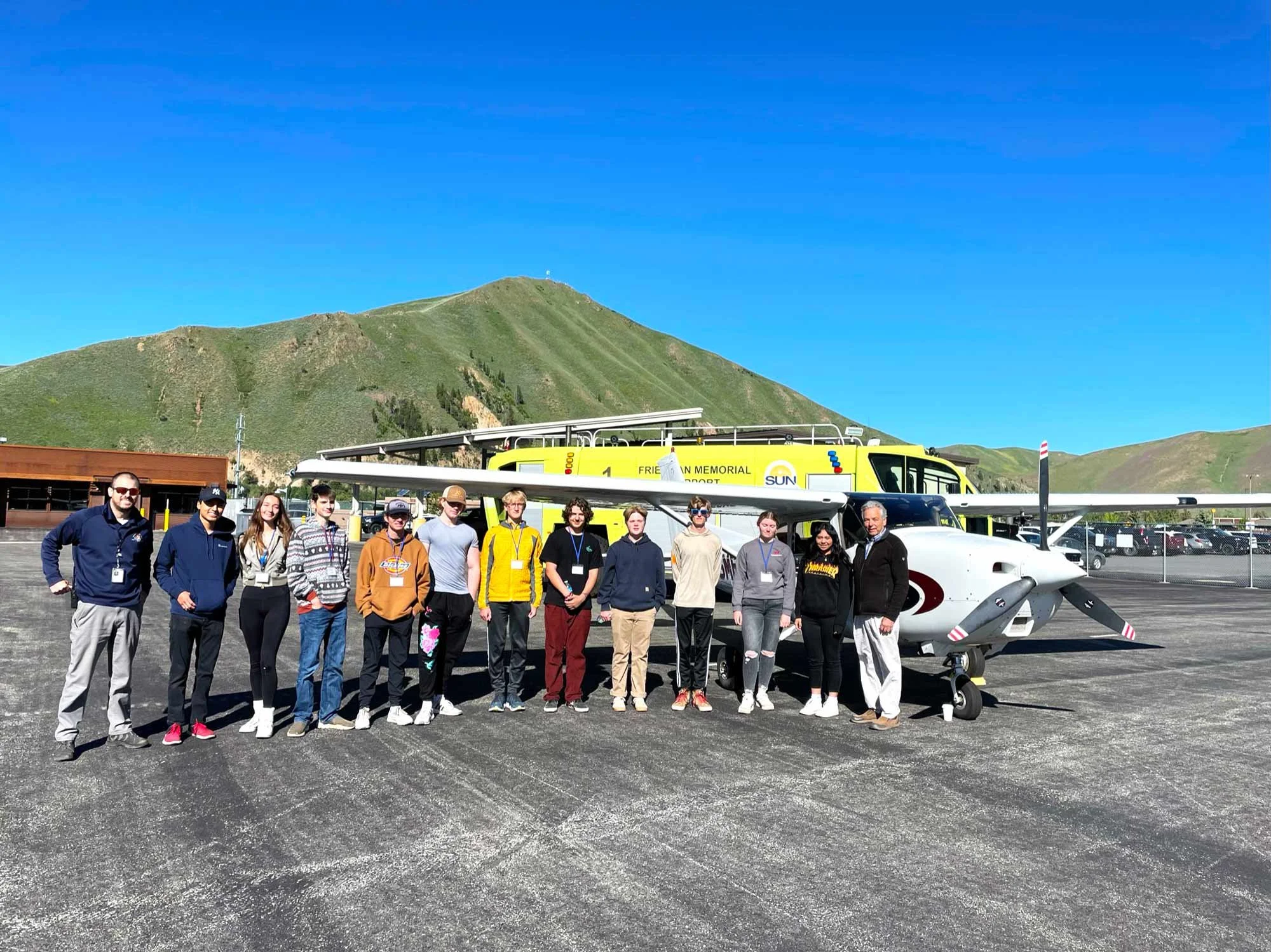 Group of people standing in front of a rescue airplane and helicopter on a tarmac, with green mountains and a blue sky in the background.