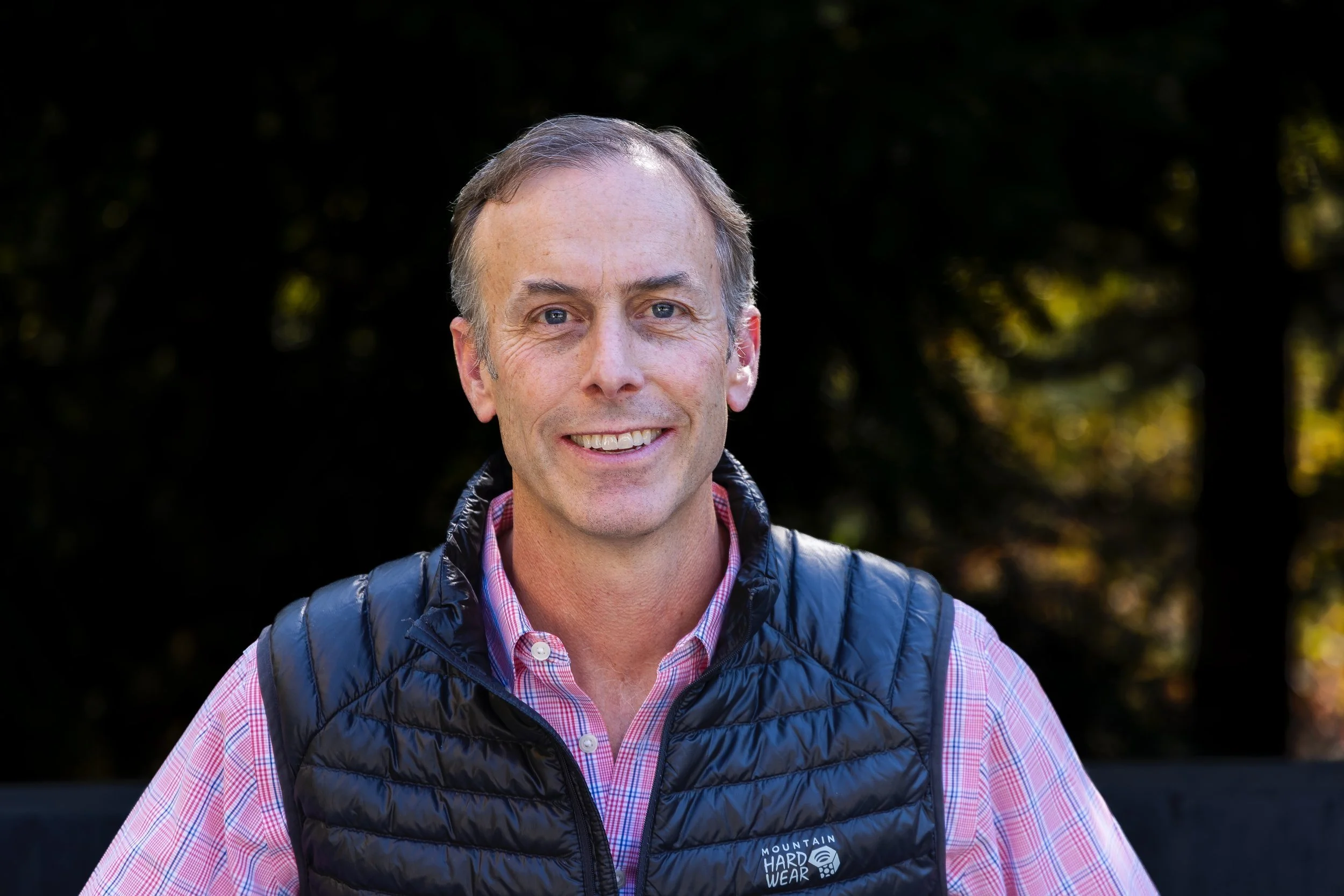 A man smiling outdoors with blurred trees in the background, wearing a pink checkered shirt and a black puffy vest.