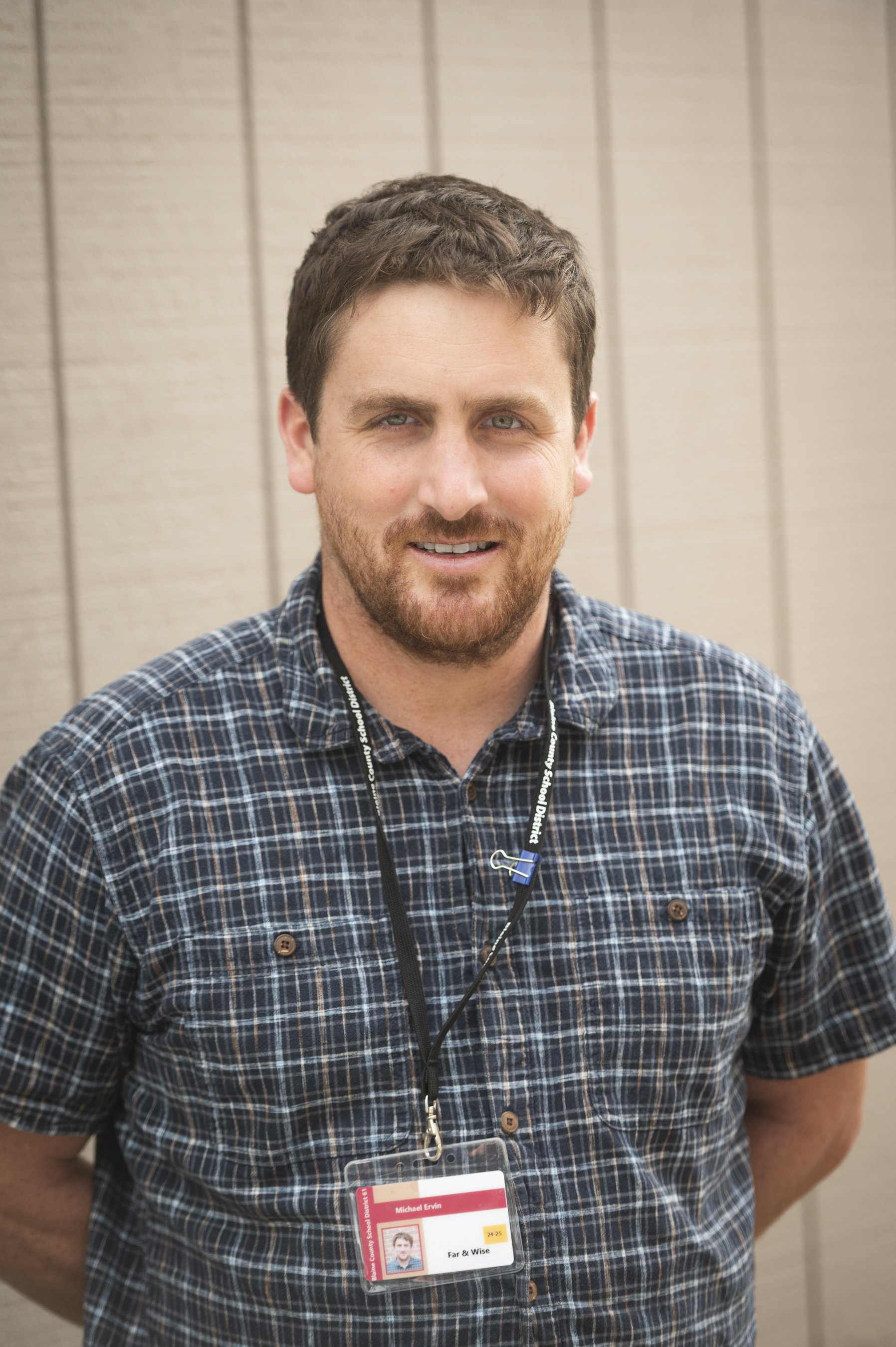Man with brown hair and beard wearing a checkered shirt and a lanyard with ID badge standing in front of a beige wall.