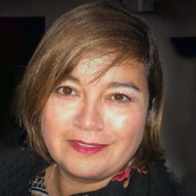A woman with short brown hair, wearing a dark top and lipstick, smiling at the camera indoors.