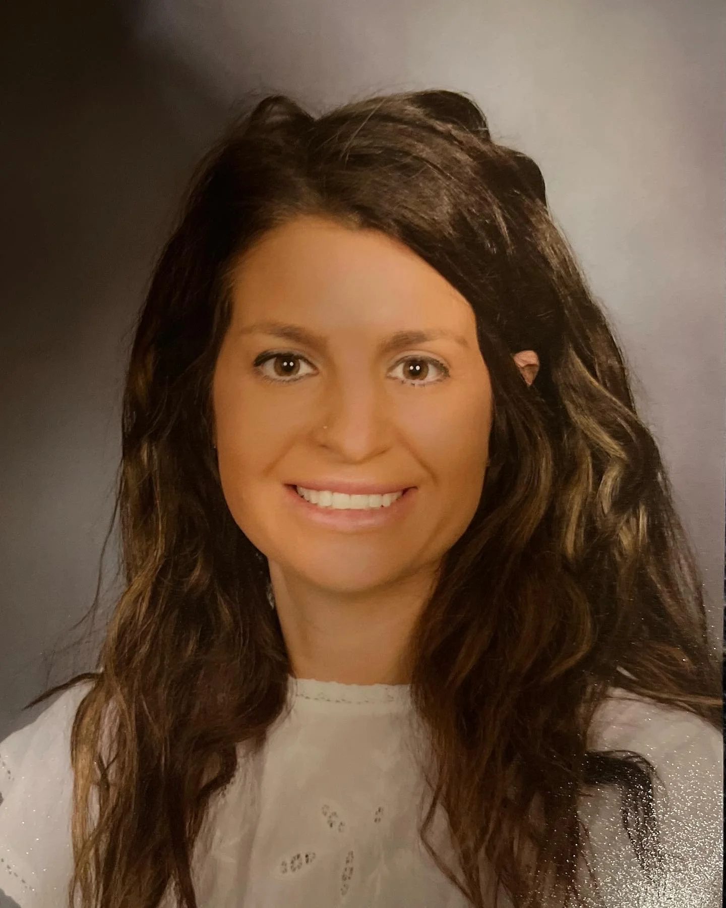 Portrait of a woman with long, wavy brown hair, smiling, wearing a white top, against a plain background.