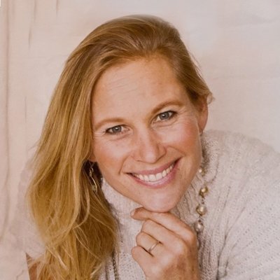 A smiling woman with long blonde hair, wearing a light-colored sweater and a pearl necklace, resting her chin on her hand.