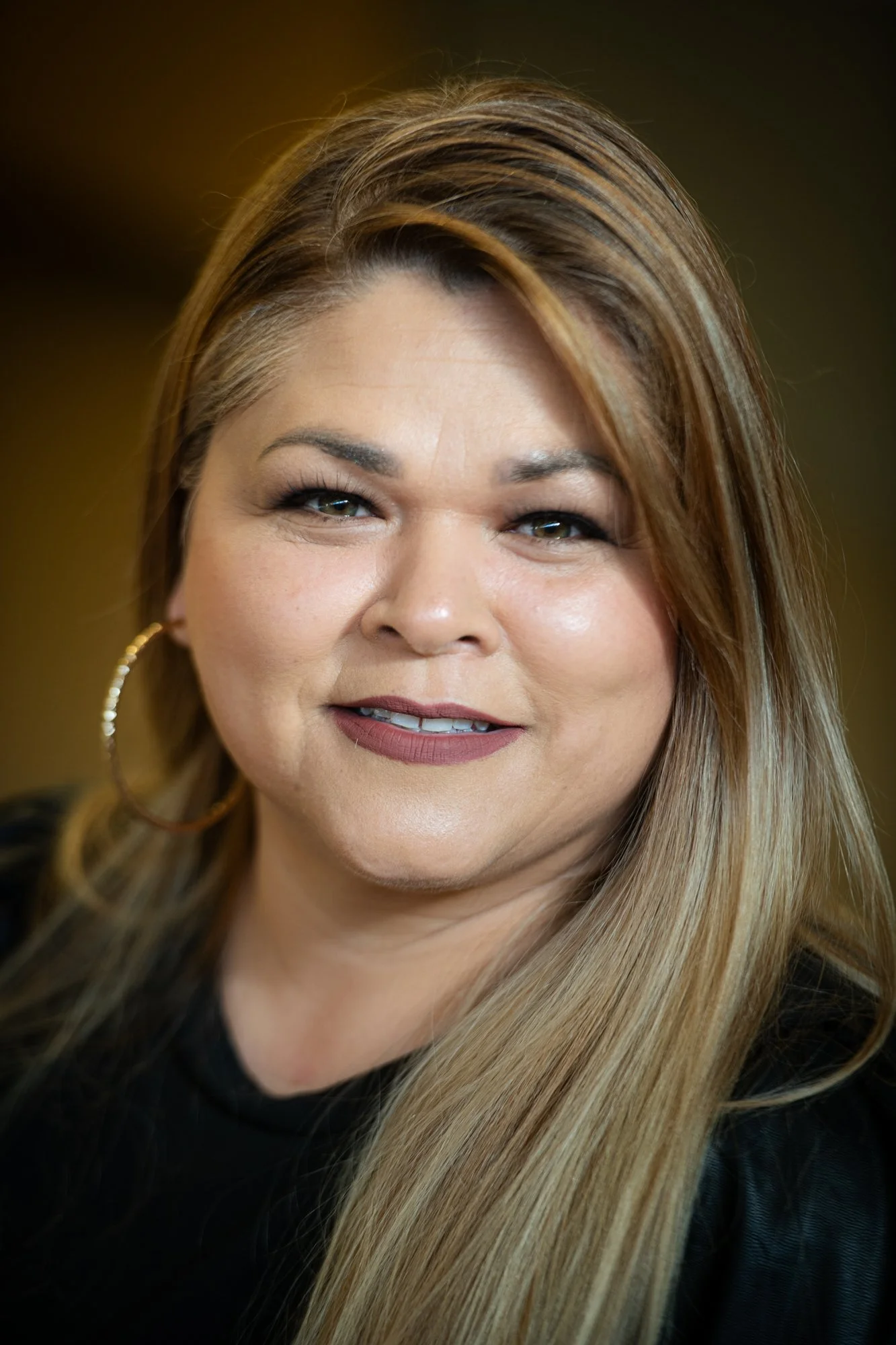 Close-up portrait of a woman with long, blonde hair and hoop earrings, smiling slightly, against a blurred warm background.