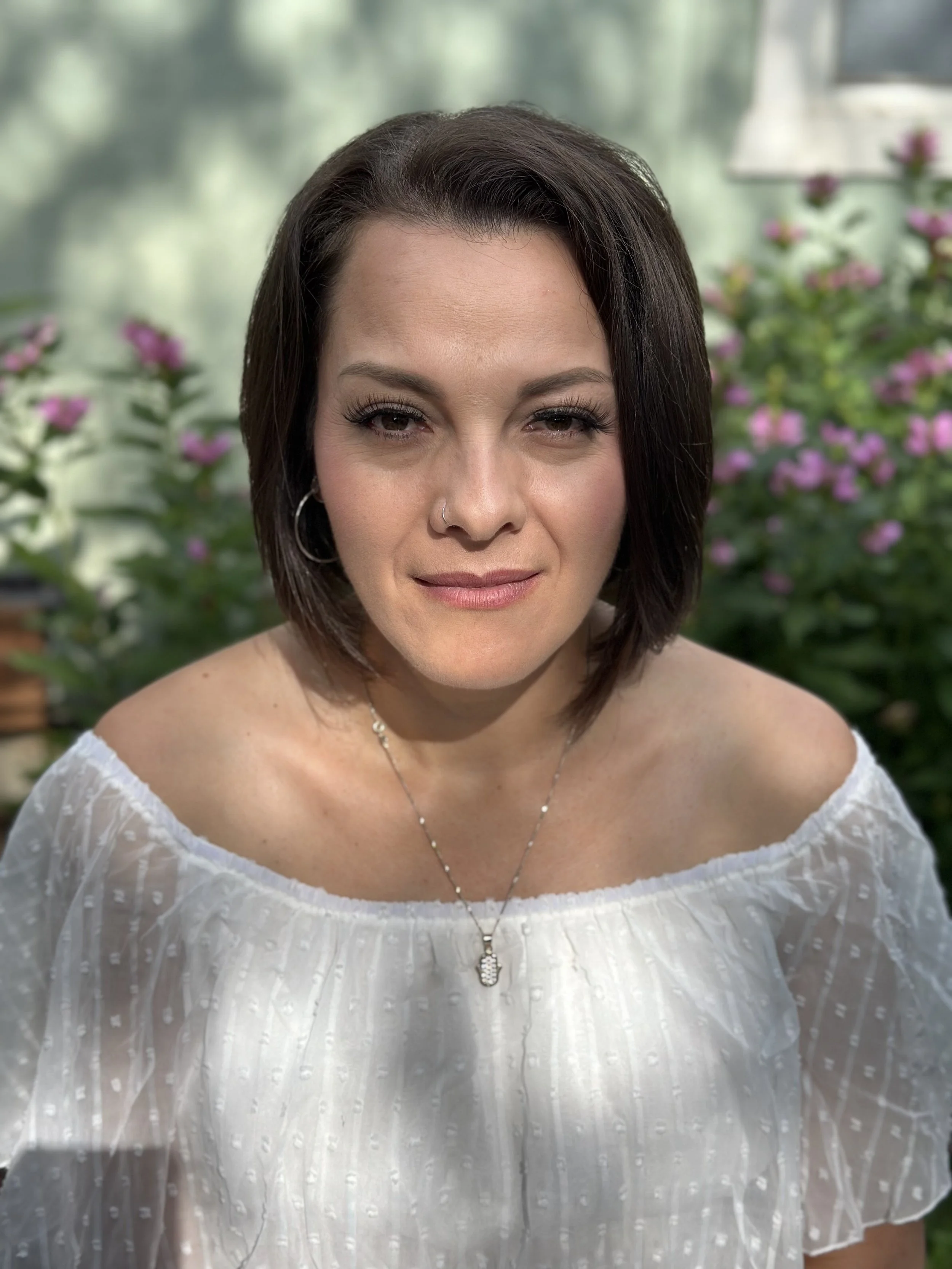 Close-up of a woman with shoulder-length dark hair, wearing a white off-shoulder blouse and a necklace, sitting outdoors with pink flowers and green foliage in the background.