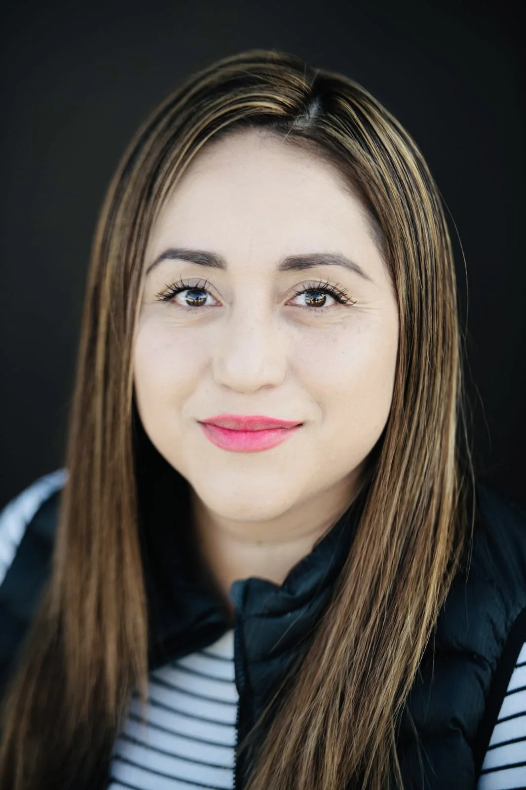 Close-up of a woman with straight, brown hair, light skin, wearing pink lipstick, a black jacket, and a striped shirt, against a black background.