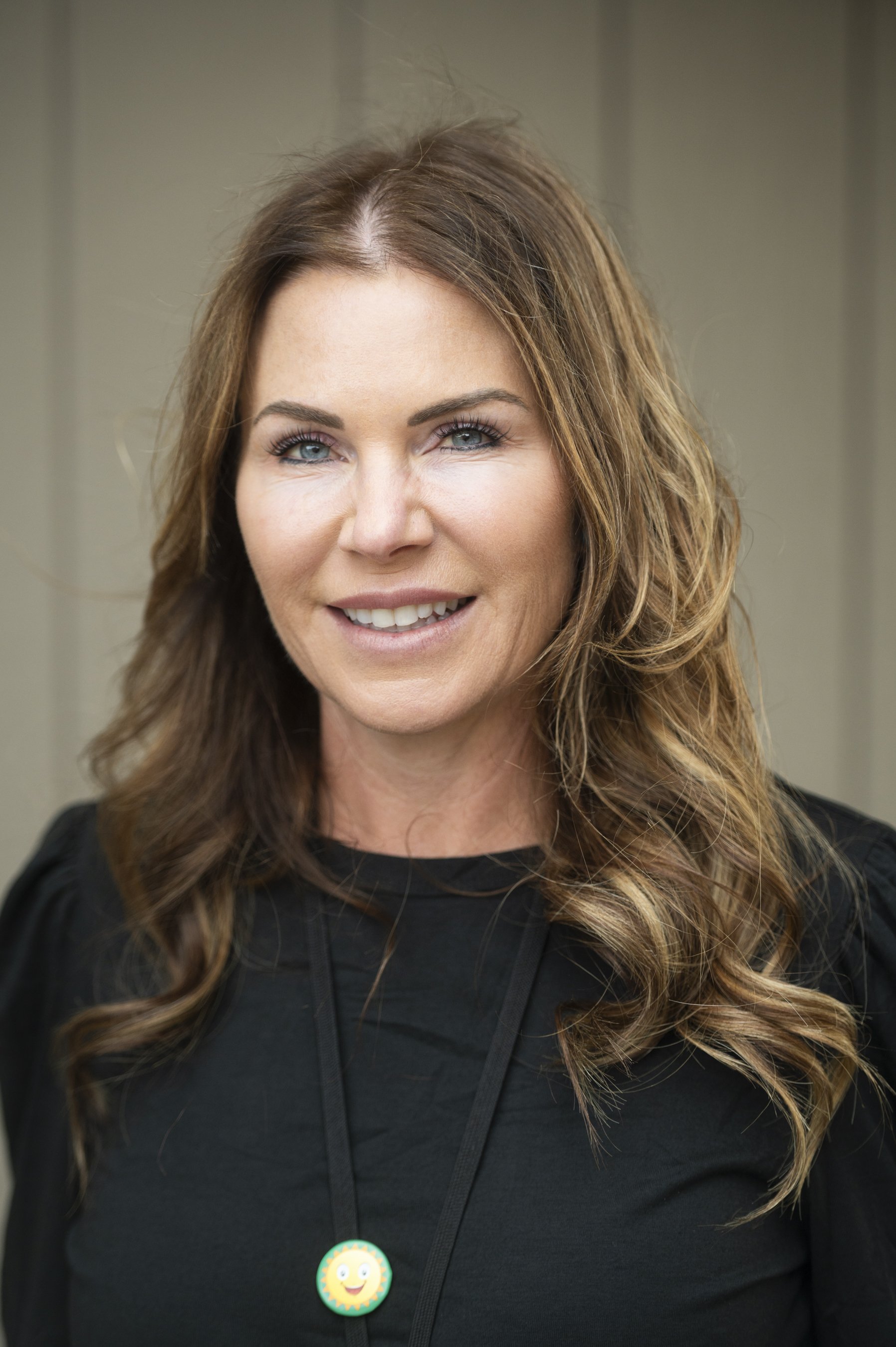 A woman with long wavy brown hair, blue eyes, and light makeup, smiling and wearing a black top with a lanyard and a sun-themed badge.