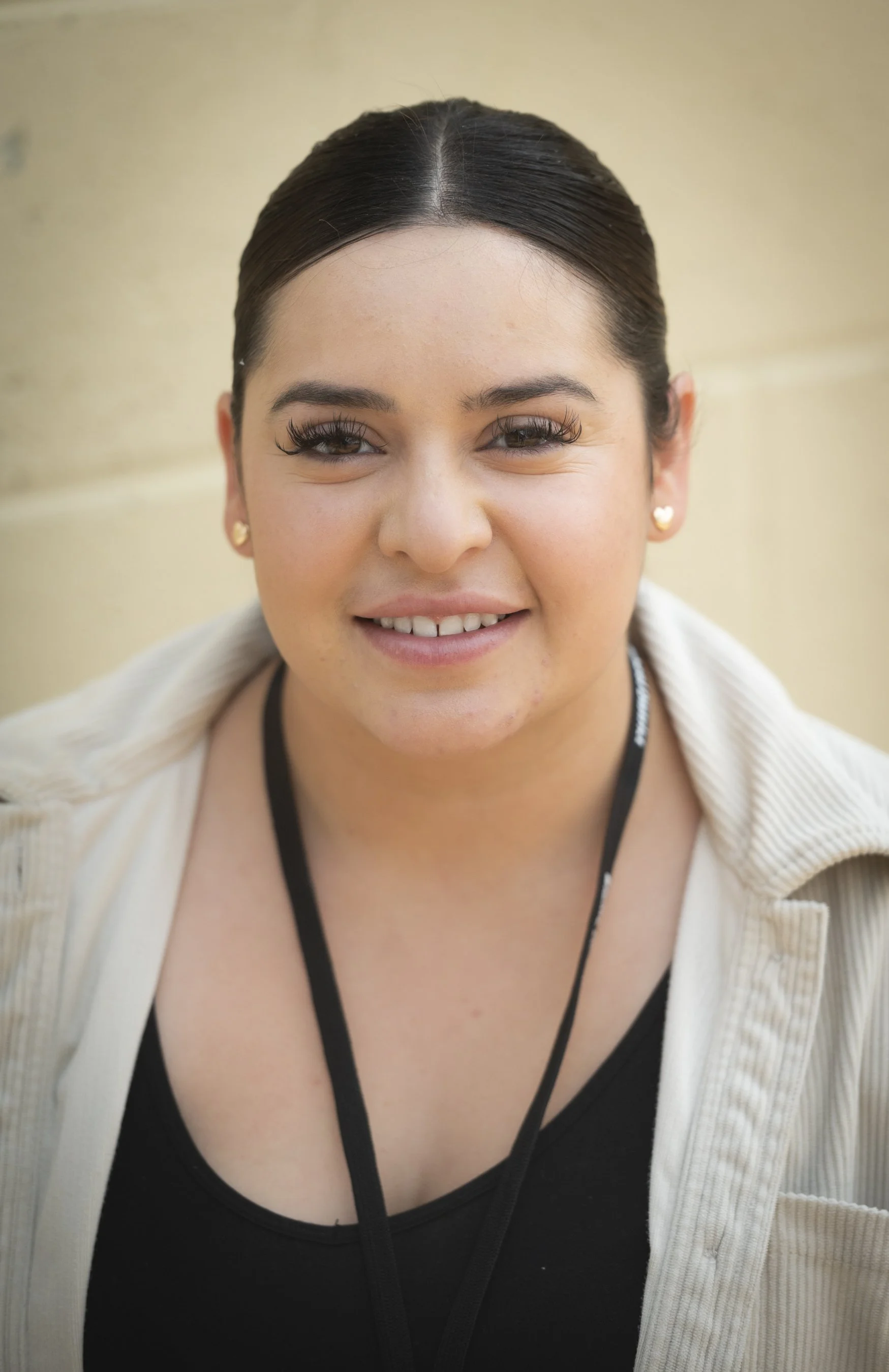 Close-up of a young woman with dark hair pulled back, wearing earrings, a black top, a beige jacket, and a lanyard around her neck, smiling outdoors.