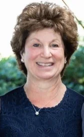 A smiling woman with short, curly brown hair and a navy blue top, standing outdoors with a background of greenery.