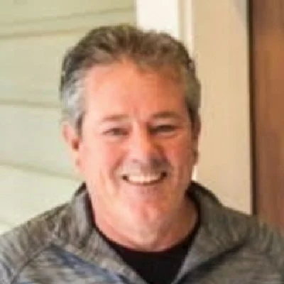 A smiling man with gray hair and a light beard, wearing a patterned zip-up sweater, indoors near a beige wall.
