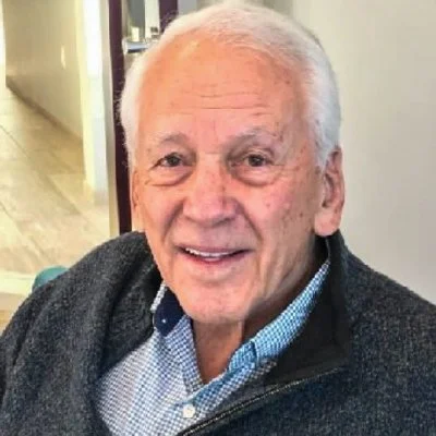 A smiling elderly man with white hair, wearing a blue checkered shirt and a dark gray jacket, sitting indoors.
