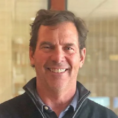 Smiling man with dark hair wearing a collared shirt and a zip-up jacket indoors.