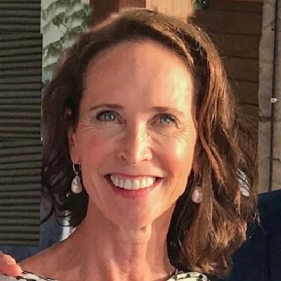 A woman with shoulder-length brown hair, smiling, wearing pearl earrings, and dressed in a black and white top, outdoors in front of a wooden fence and greenery.