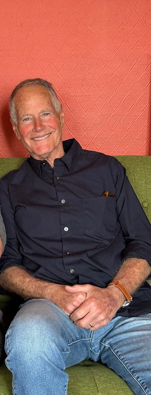 An elderly man sitting on a green sofa, smiling, wearing a dark button-up shirt and light blue jeans, against a textured reddish-orange wall.