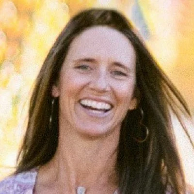 A woman with long brown hair smiling outdoors with colorful autumn trees in the background.