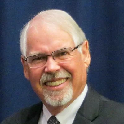 Portrait of a smiling older man with glasses, gray hair, and a beard, wearing a suit and tie.