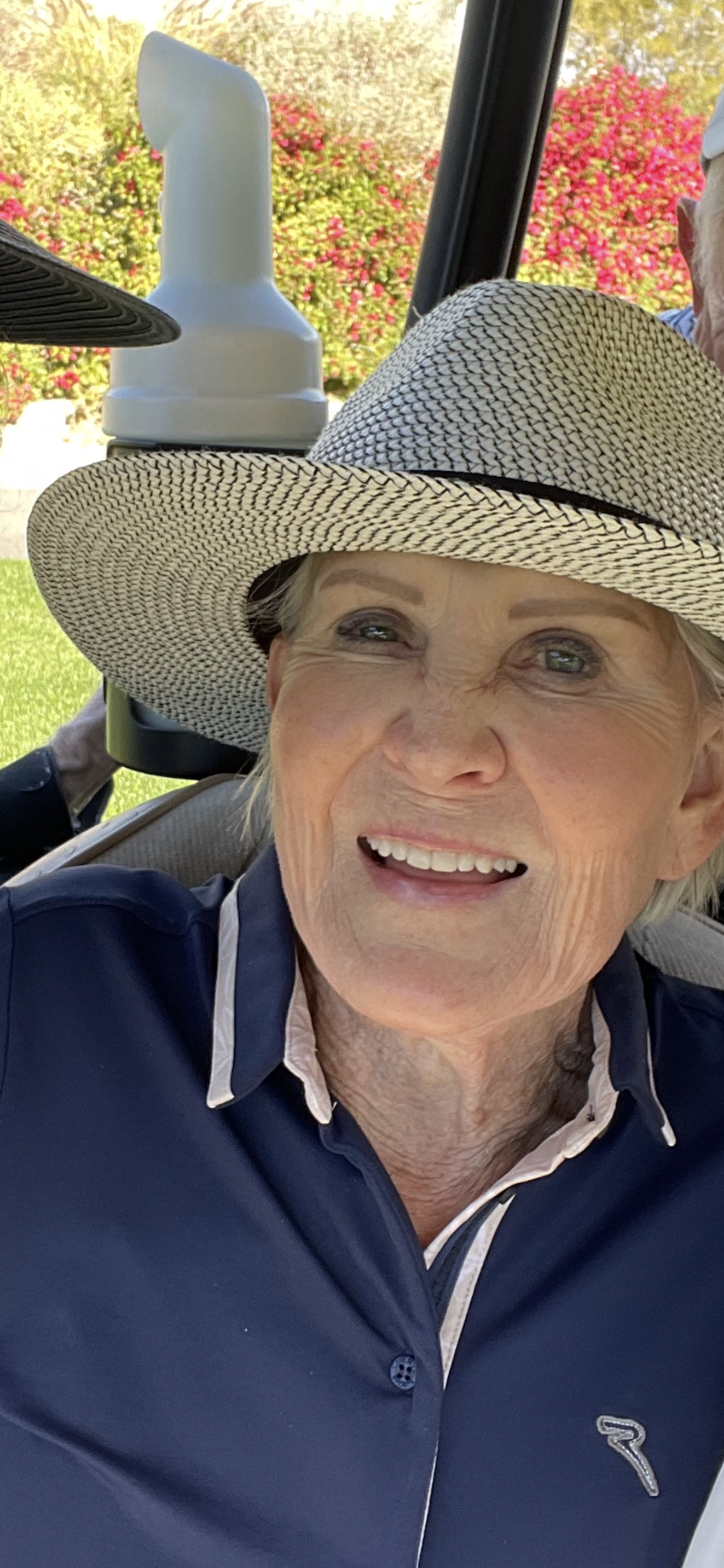 Smiling elderly woman wearing a wide-brimmed hat and a navy blue collared shirt, outdoors with colorful bushes in the background.