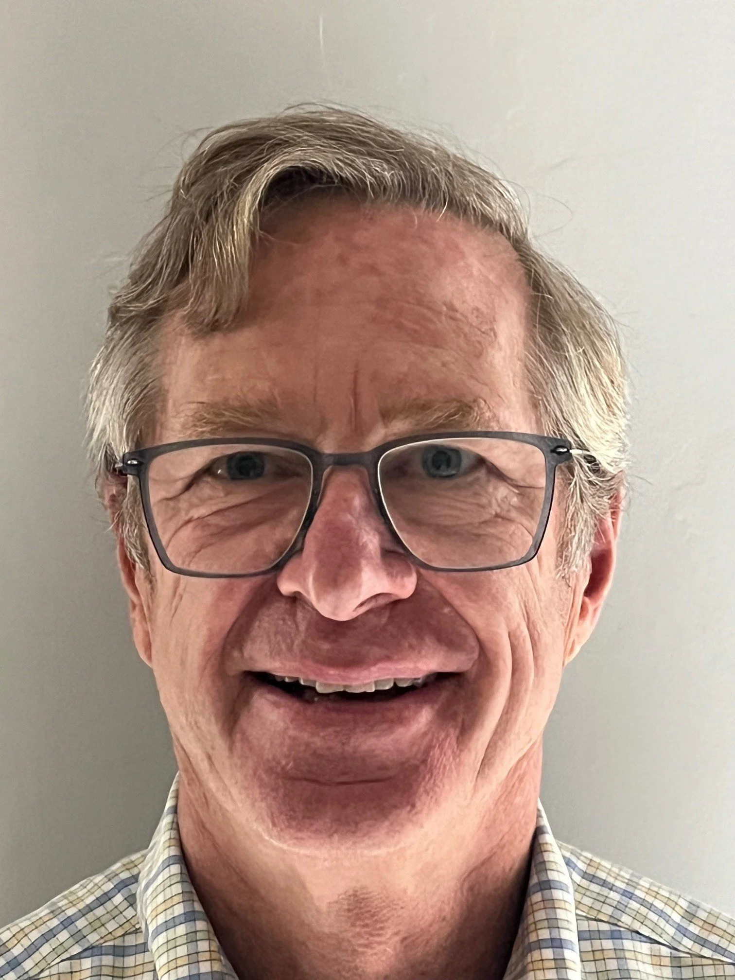 A smiling middle-aged man with light-colored hair, wearing glasses and a checked shirt, in front of a plain wall.