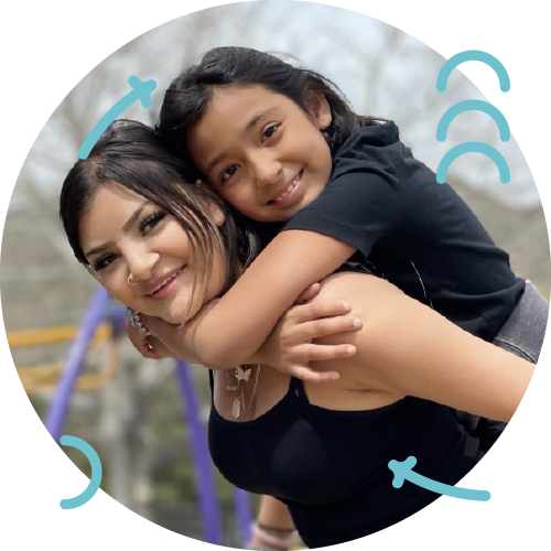 A woman and a young girl smiling, with the girl hugging the woman from behind at a playground.