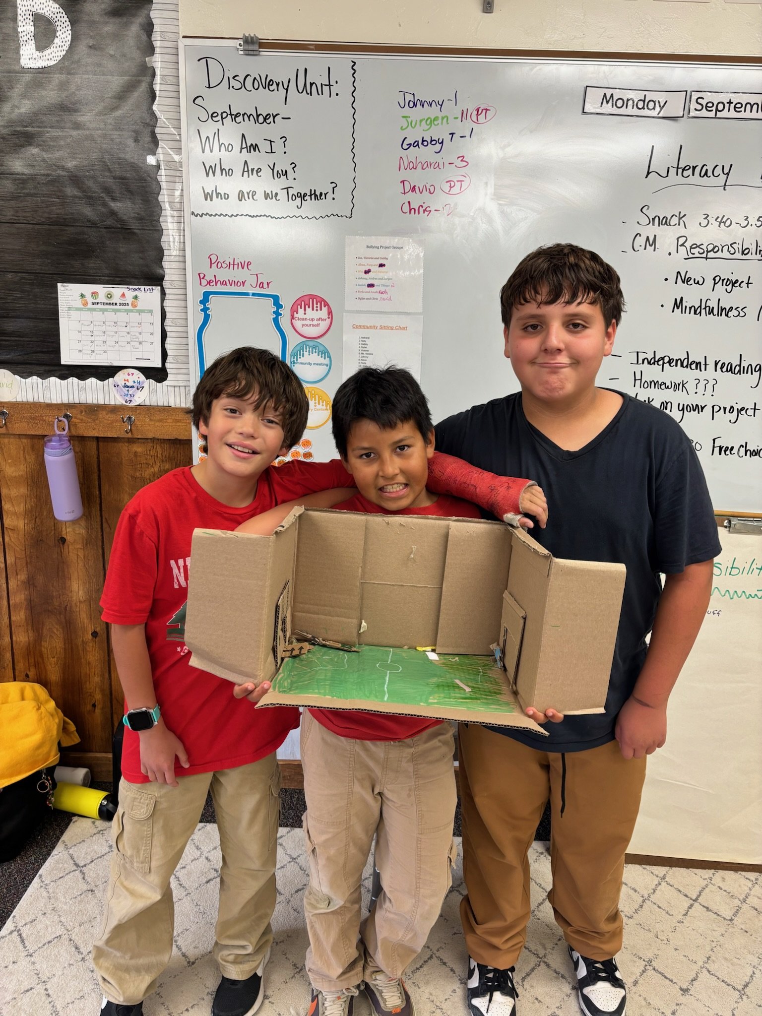Three boys in a classroom holding a small cardboard soccer field model with a green playing surface. Behind them are a whiteboard with notes and a wall with a calendar and a motivational poster.