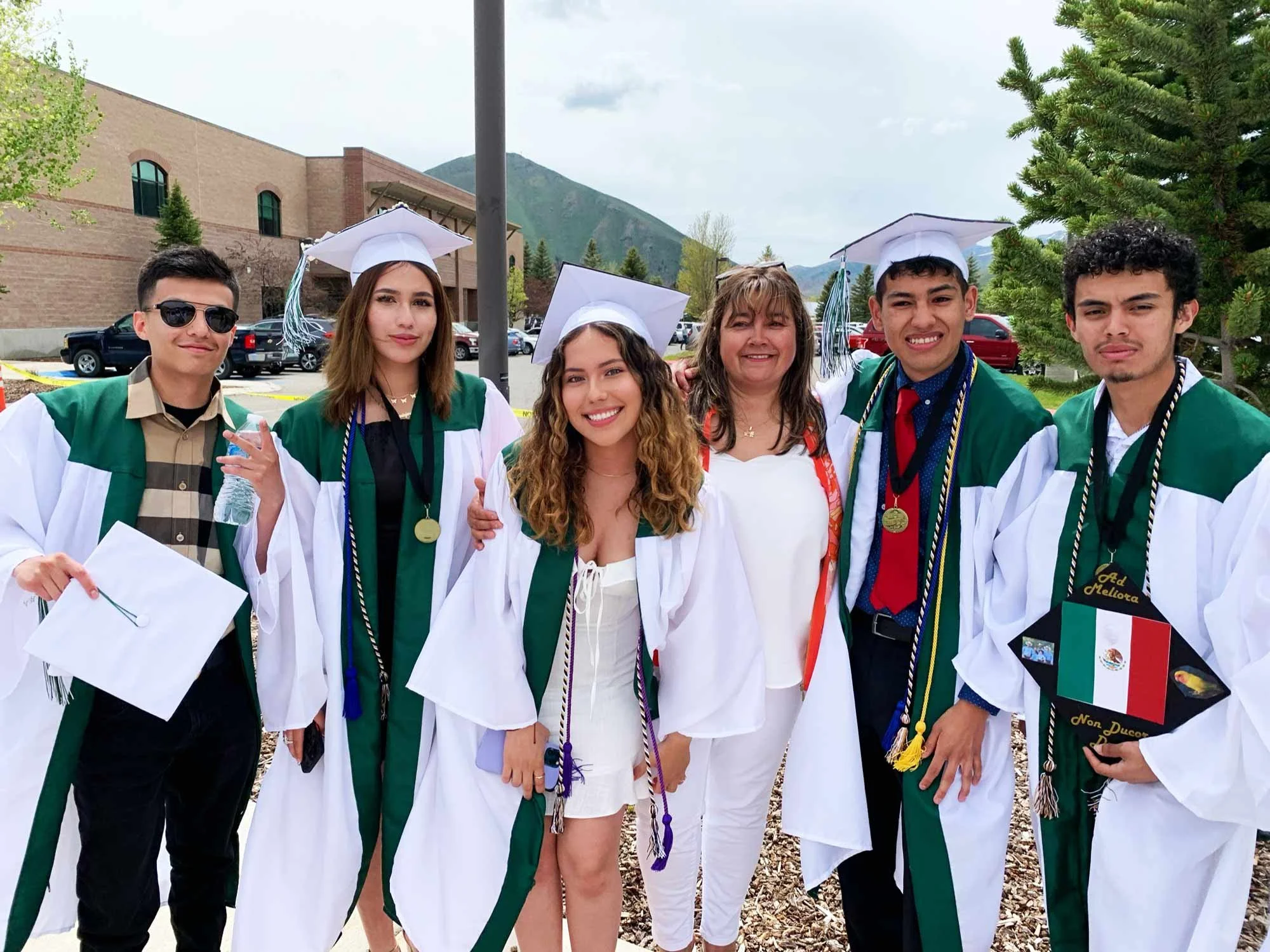 Group of six diverse graduation students and their teacher outdoors, wearing caps and gowns, with some holding diplomas and medals, smiling, mountains and a building in the background.